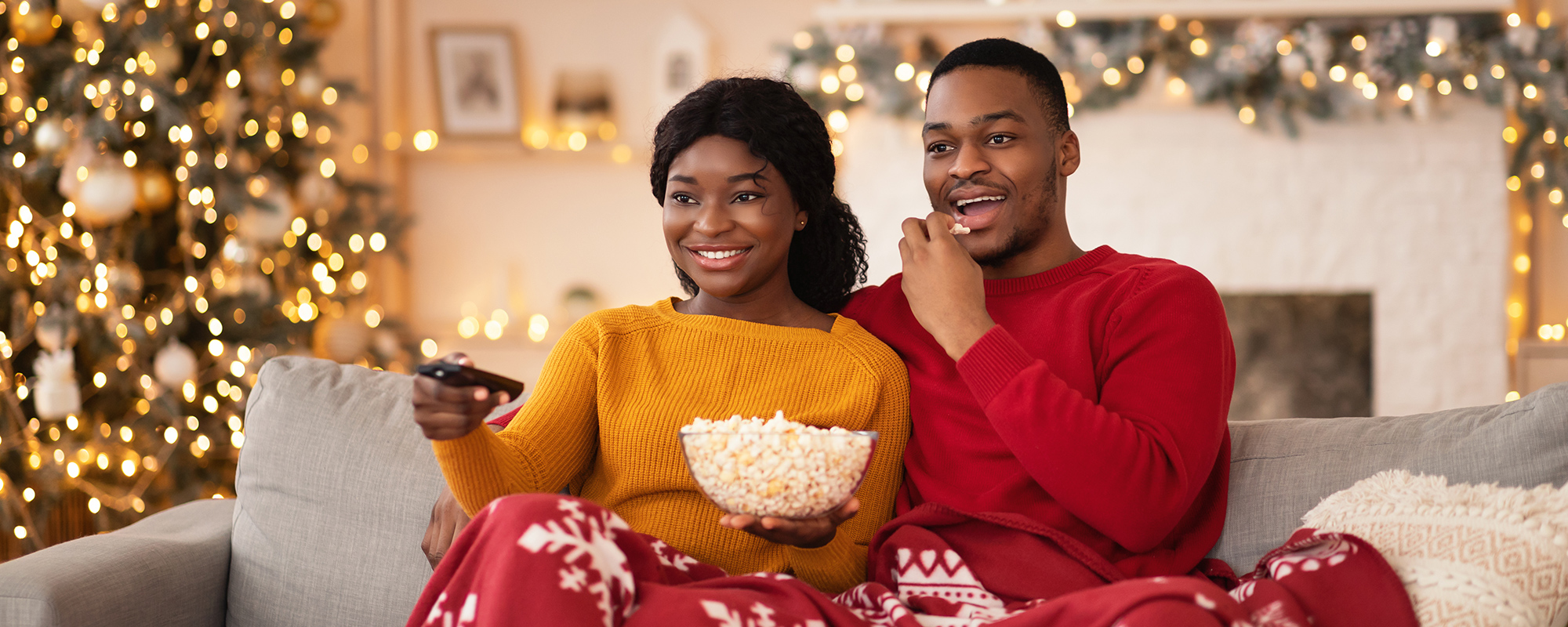 A happy couple sitting on a safe with a Chrstmas blanket across their laps. She;s pointing the remote at the TV and he's eating some popcorn from the big bowl they're holding