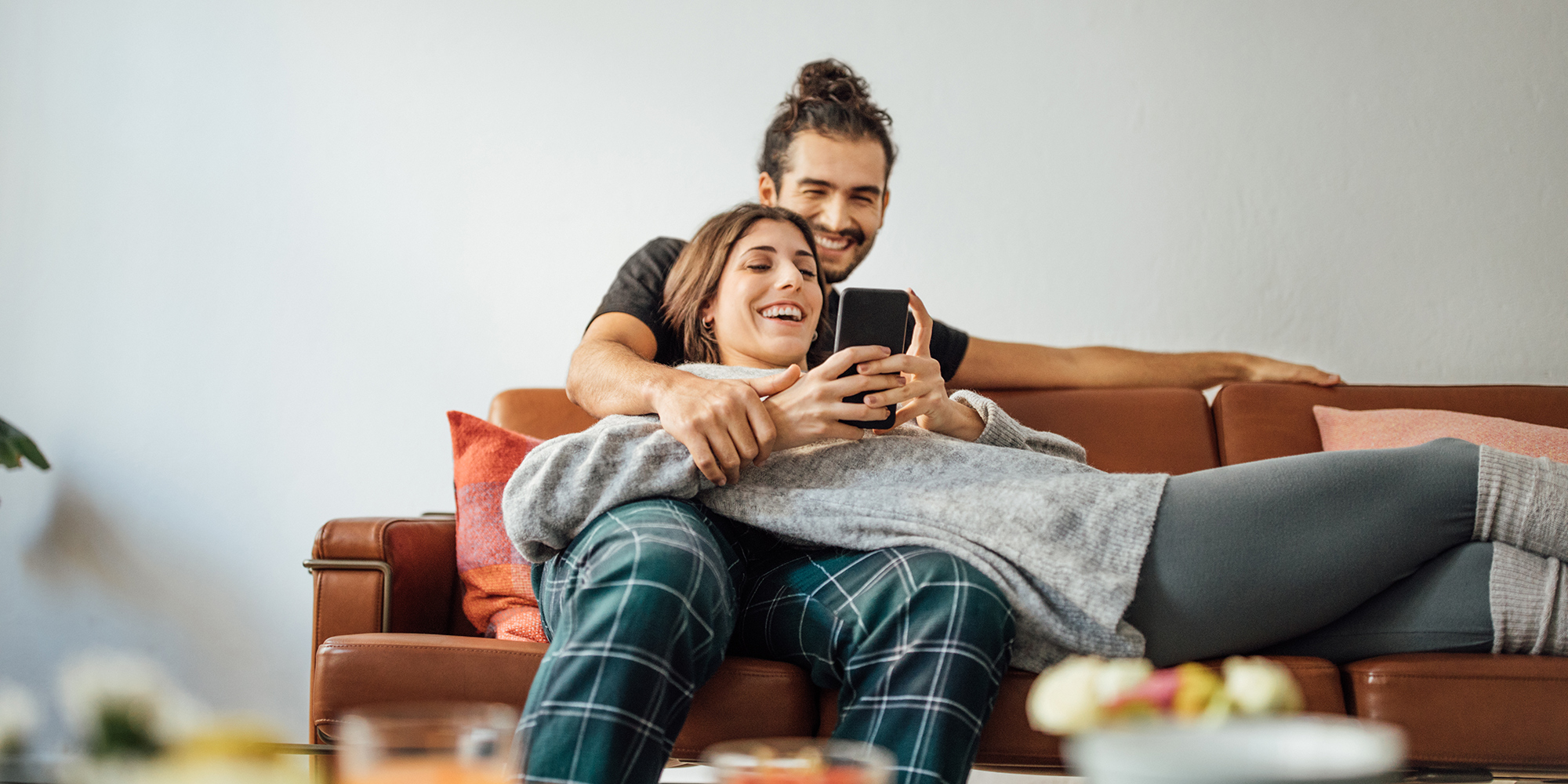 A young couple laying on a sofa smiling at the smartphone one of them is holding.