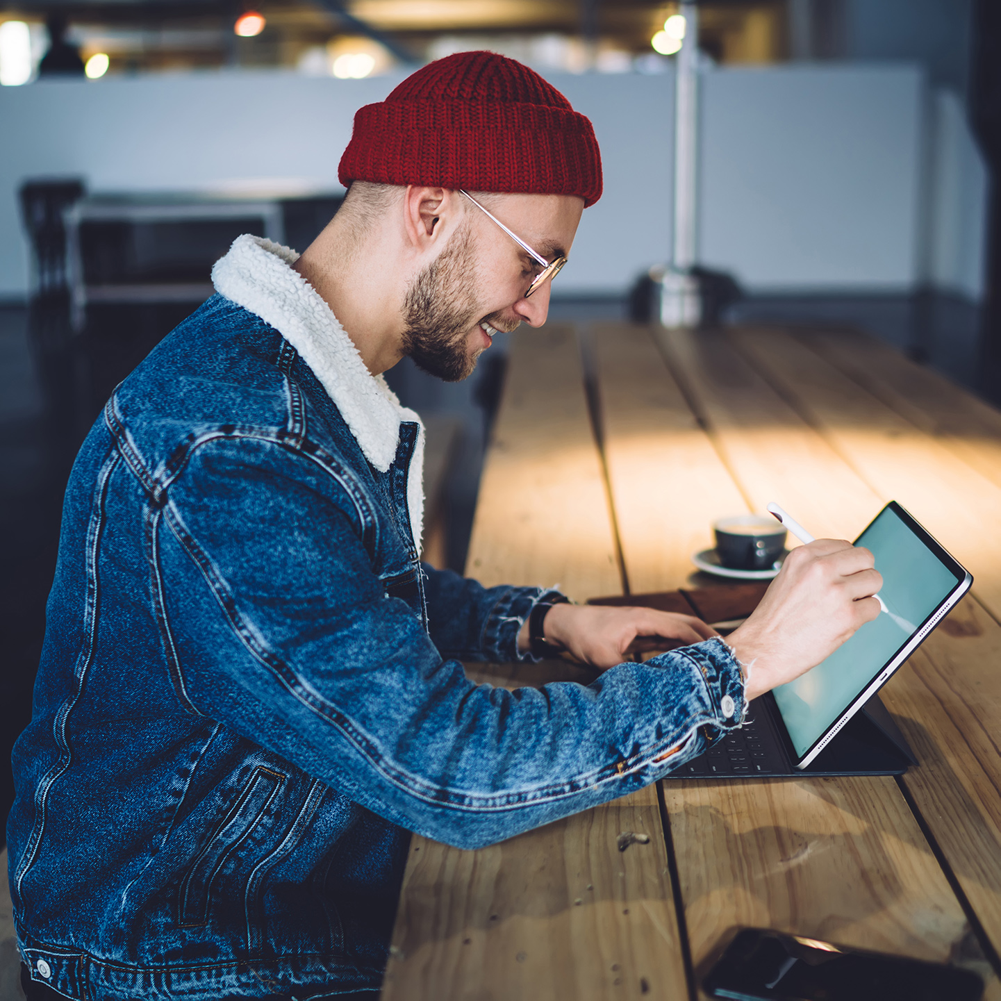 Man sitting at a very long table. He's smiling at his tablet device and using the touchscreen of it with his hand.