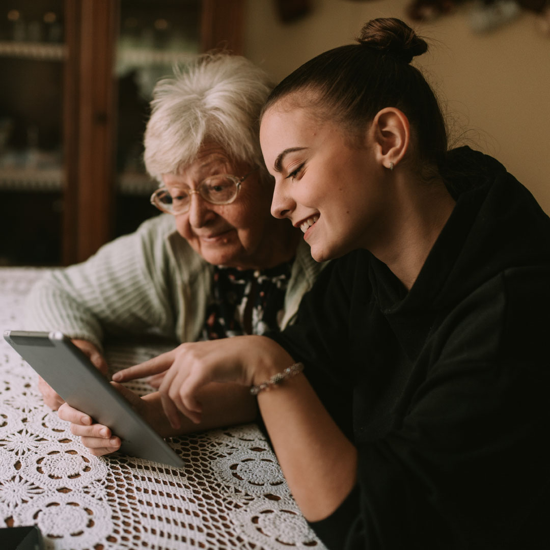 Oler lady and younger woman sitting together smiling. The younger woman is pointing at something on her tablet.