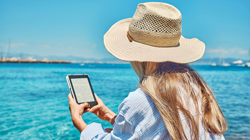 Person sat by the sea, reading a book on an e-reader