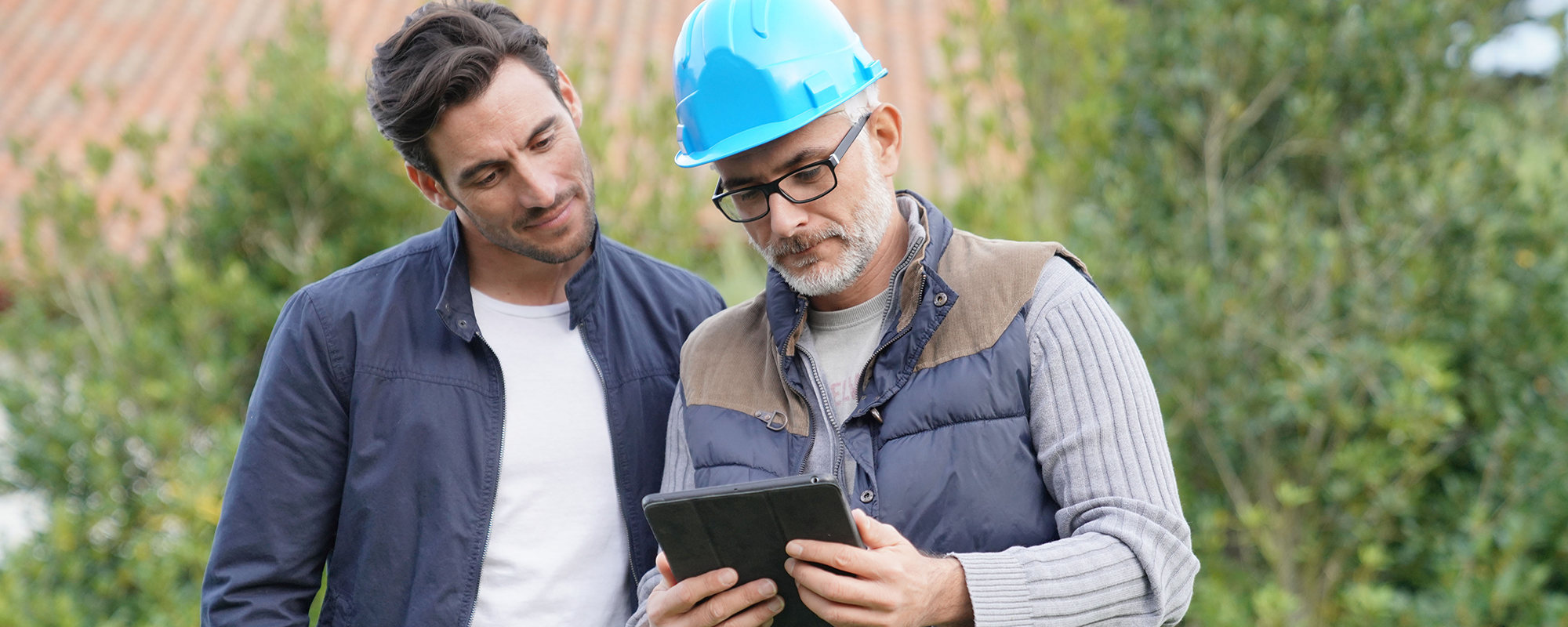 two men standing outside looking at a tablet, one of the men is wearing a blue hard hat.