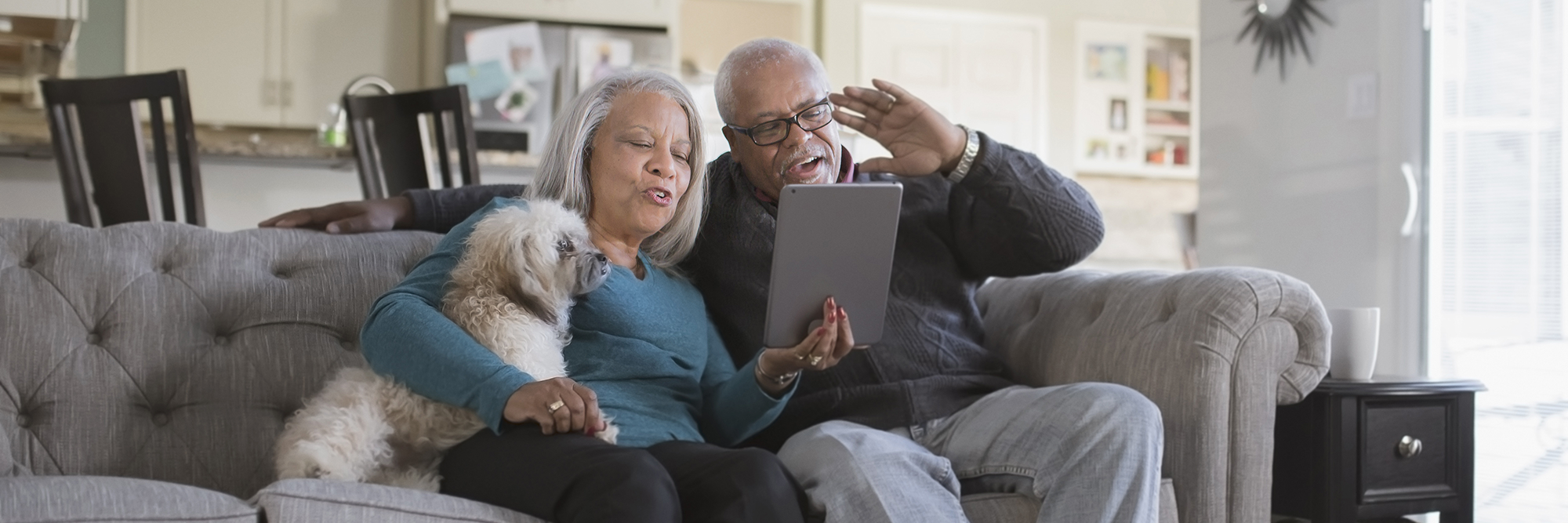 an older man and woman sitting on the sofa with a dog. They are having a video chat with someone on their tablet.