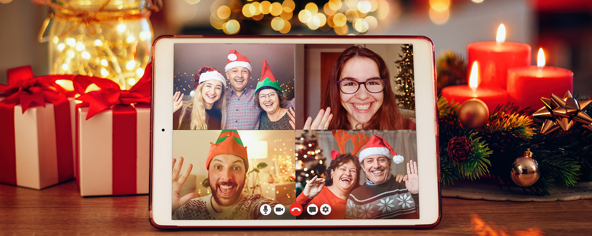 This image shows a picture of a tablet being propped up among some Christmas decorations on a table being used for a video call with four images of people waving at the person using the device. They're all very happy and festive.