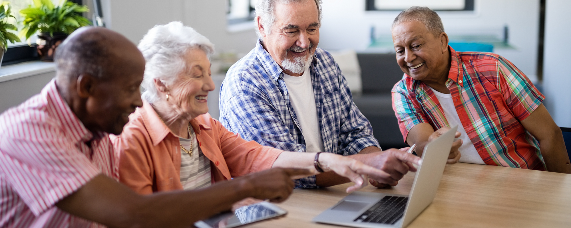 four older people sitting at a table looking at a shared laptop. They're all smiling.