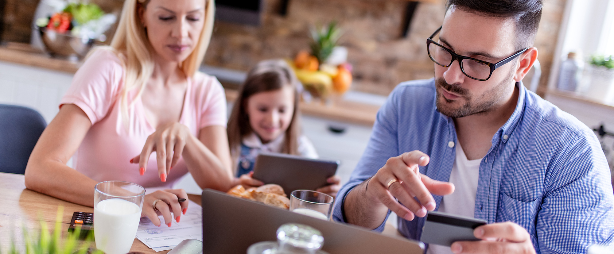 young couple in a kitchen doing some paperwork at the table whilst their young daughter is looking at her tablet  | Digital Wings | understand the cost of living