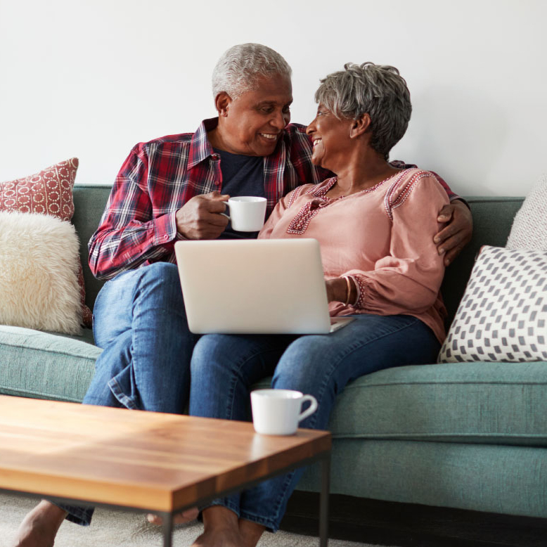 older man and woman sitting closely together on a green sofa. The lady has a laptop on her lap, and he's holding a mug in his hand. They're smiling broadly at each other.