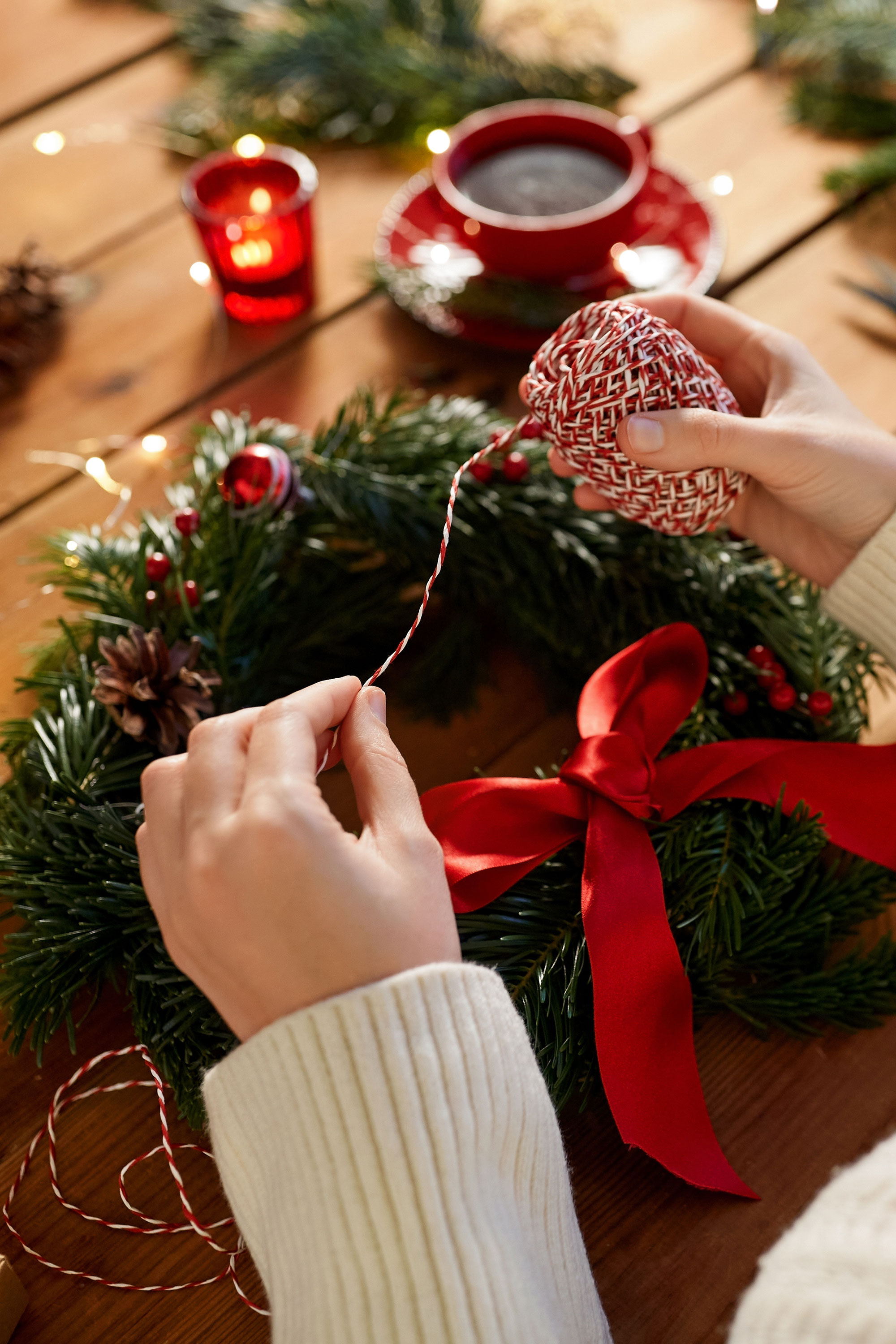 This image shows the hands of someone creating a homemade Christmas wreath which has pinecones, red and white twine and a big red satin bow.
