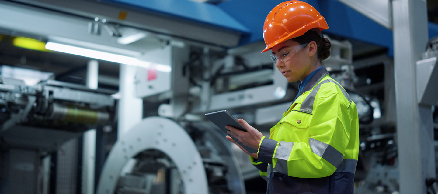 woman wearing a yellow high vis jacket and an orange hard hat in an industrial environment. She's looking at her tablet in her hand concentrating.