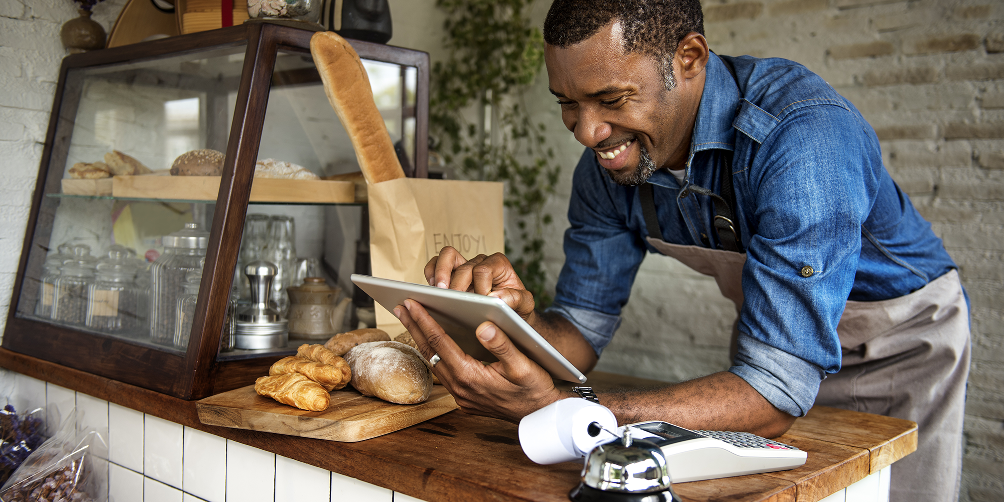 Black man behind the counter of a cafe smiling whilst using a tablet device