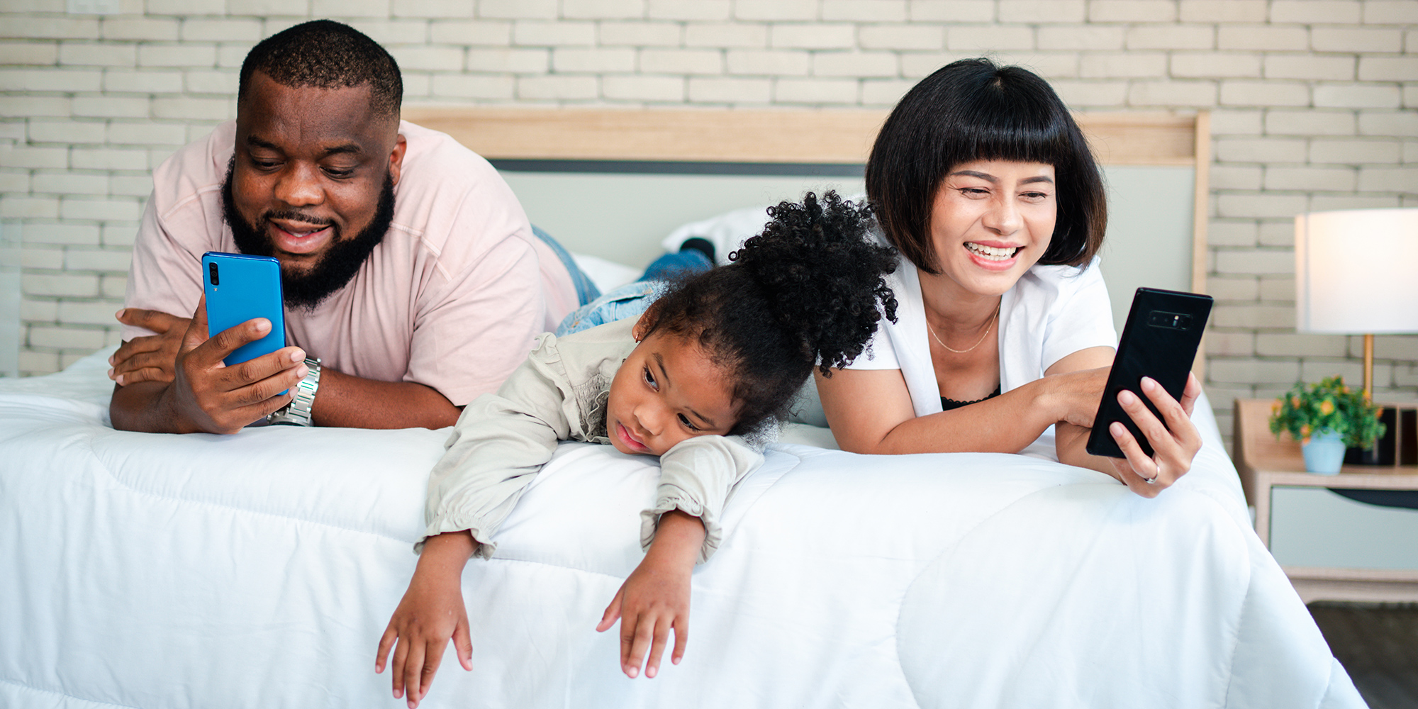 mother and father laying on a bed using their mobile phones while their child looks bored and ignored between them | Digital Wings What is Digital Wellbeing