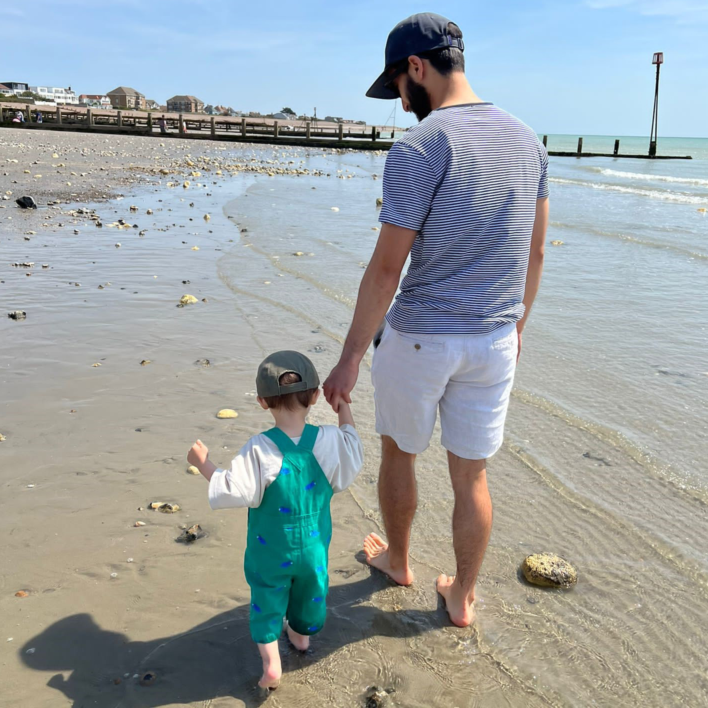 Digital Eagle Matt Song holding hands with his toddler, they're walking along the beach on a sunny day away from the person taking a photo. Matt is looking down at his child .