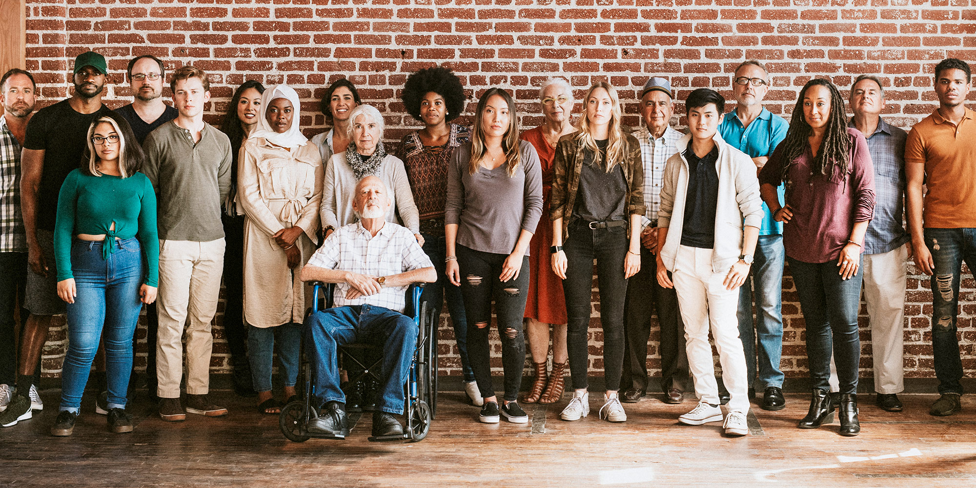 group of people of various ages, ethnicities and abilities standing/sitting in front of a red brick wall