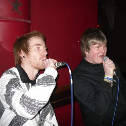 Digital Eacle Mike Haigh and his friend are singing Karaoke together, both are holding a microphone each standing on a stage with red walls behind them.