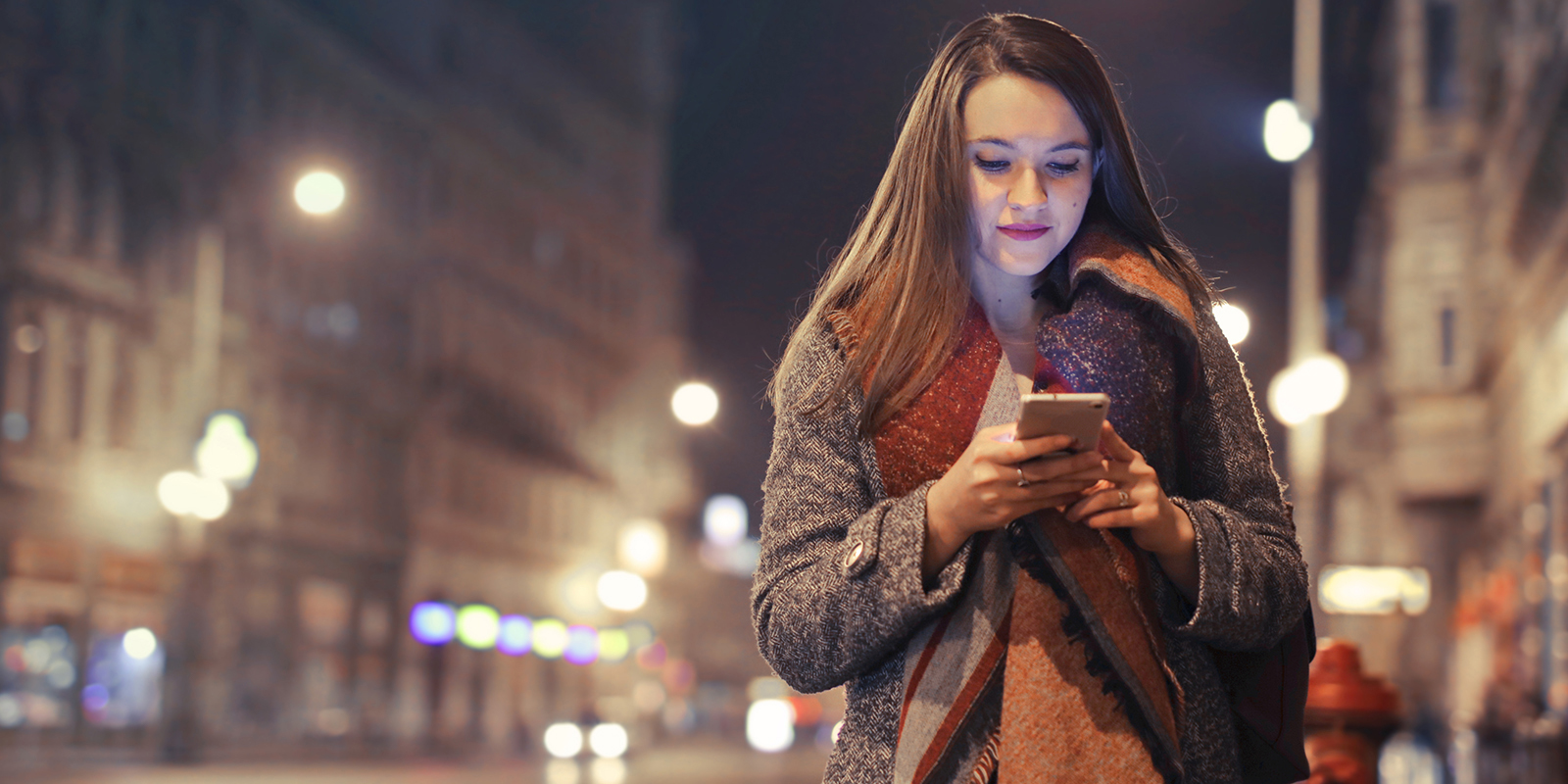 young woman walking down a well lit high street at night time, she'w wearing a winter coat and she's looking at the smartphone in her hand.