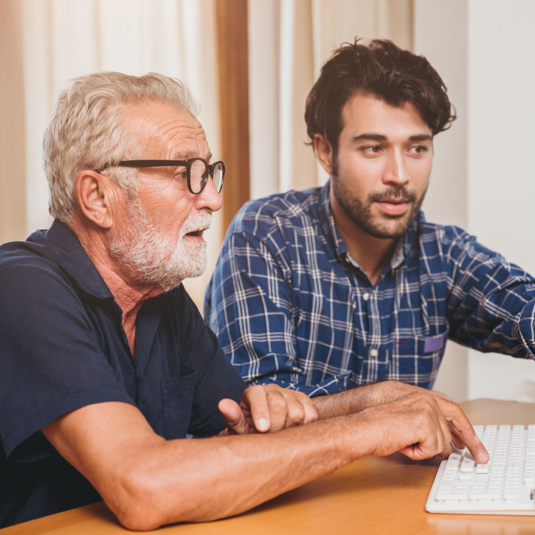 older man and younger man sitting at a table next to each other. The older man is typing on a computer keyboard whilst the younger man is pointing at the screen which they're both concentrating on.