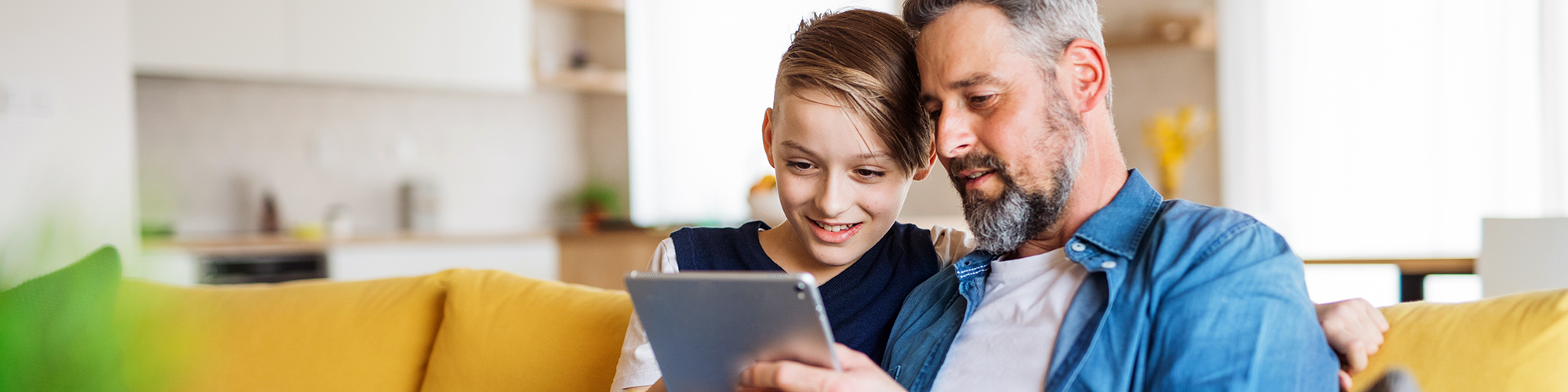 Father and son, looking at a tablet, sat on a sofa.