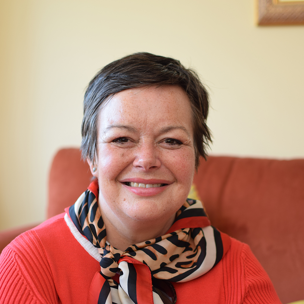 Headshot image of Paula Fowler smiling at the camera. She's wearing an orange jumper and has a leopard print silk scarf around her neck