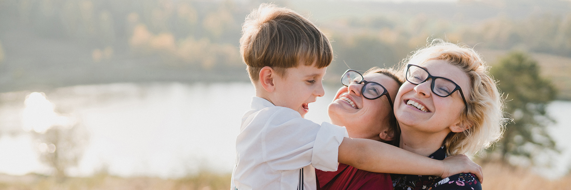 Two ladies cuddling a little boy in front of a lake all smiling with the sun shining behind them | Digital Wings What is Digital Wellbeing