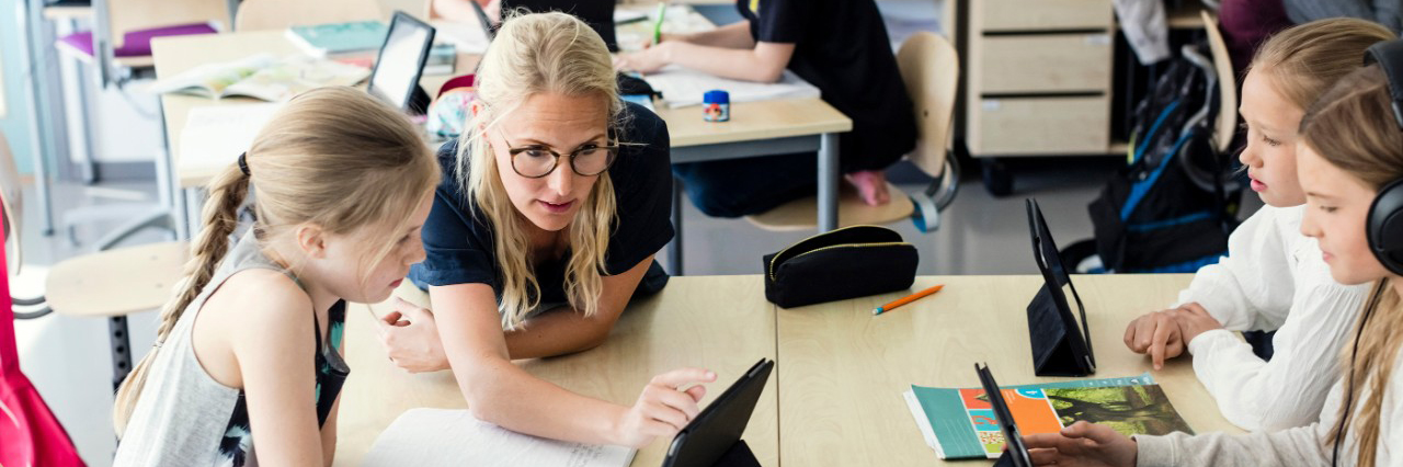 Lady sitting at a desk with children using a tablet.