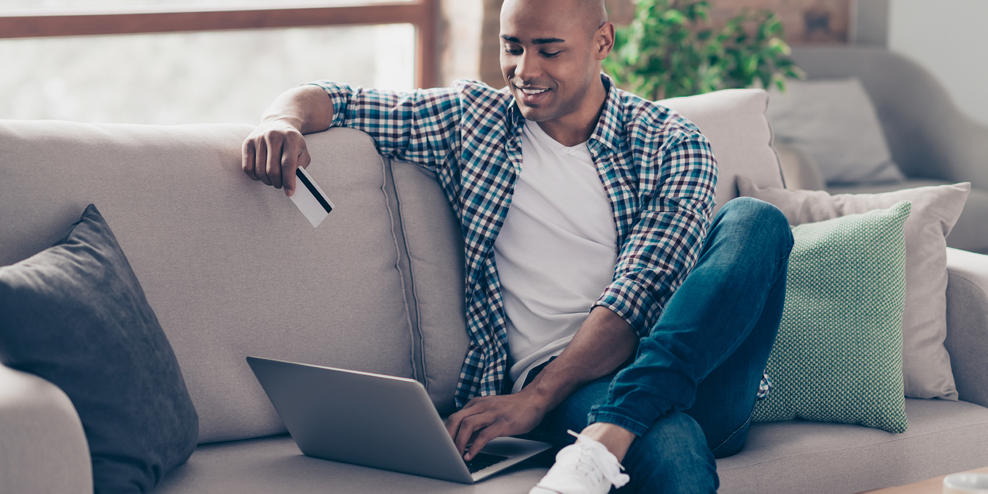 Man sitting on a sofa looking at his laptop holding a payment card | Digital Wings | Online money management