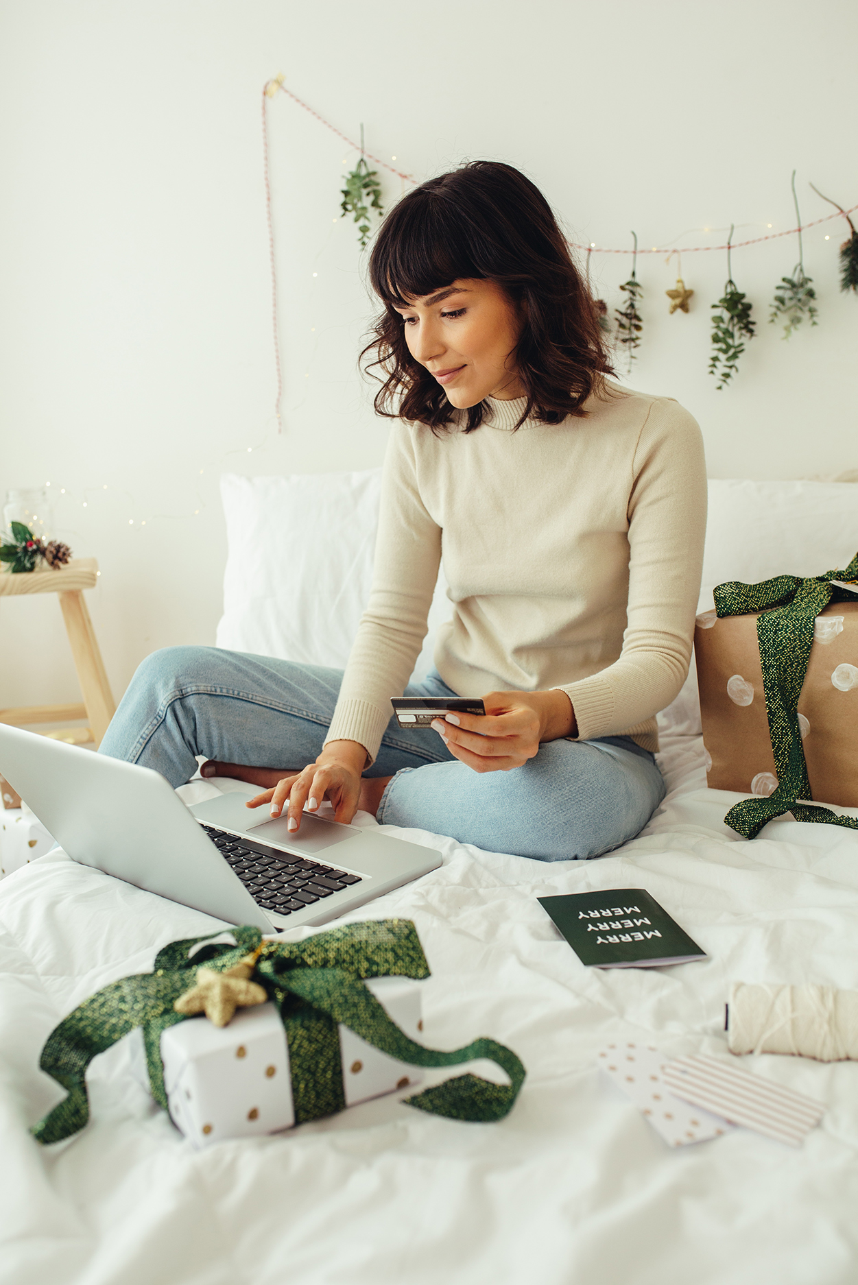 woman sittig on a bed doing some online shopping on her laptop with some beautifully wrapped gifts on the bed next to her, there are Christmas decorations on the wall.