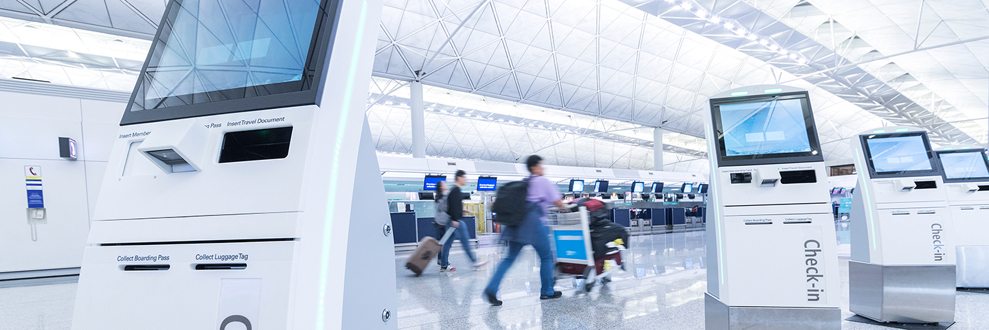 Row of self check-in desks at an airport with people with suitcases in the background.