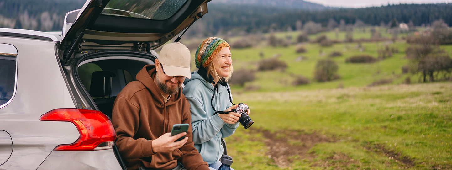 A happy looking couple sat in the open boot of their car in the middle of the countryside. She's holding a camera and he's looking at his smartphone.