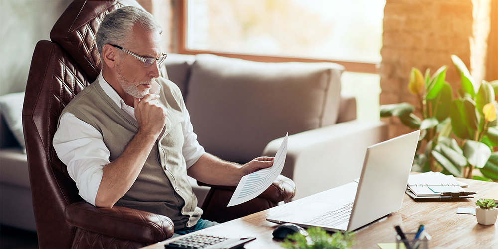 older man looking at a sheet of paper in front of a laptop