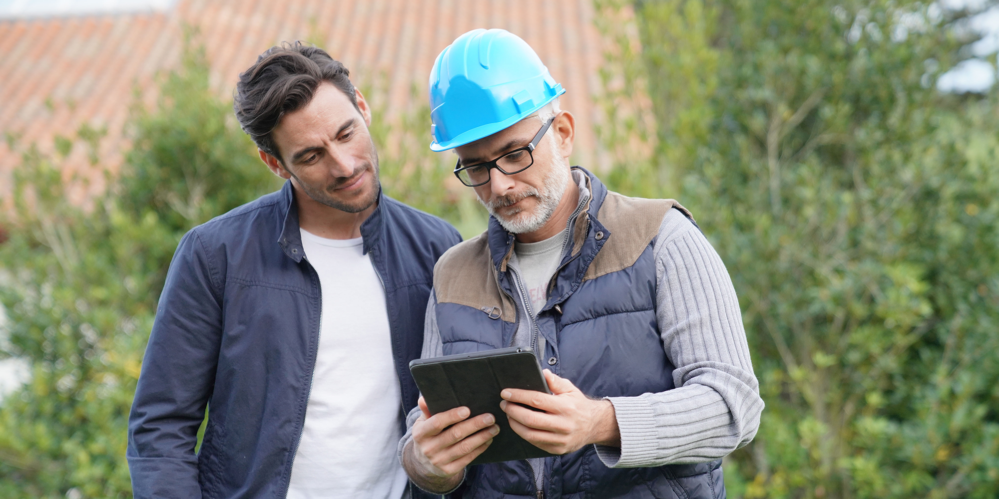 younger man next to an older man wearing a hard hat looking at a tablet
