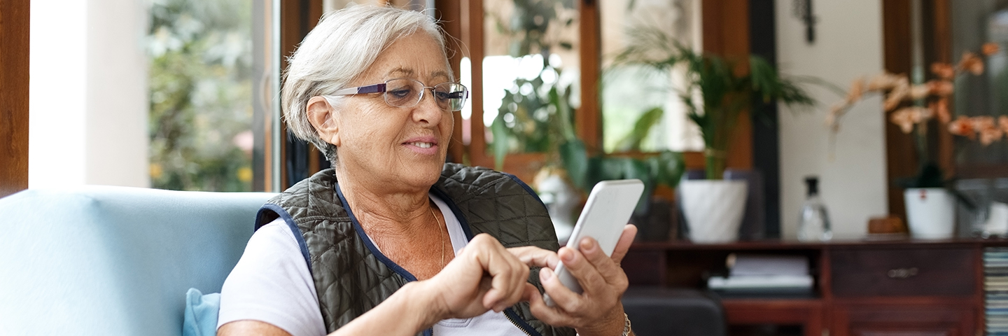 mature lady sitting on an armchair in her home smiling at her smartphone. She's using her finger to select something on the screen.