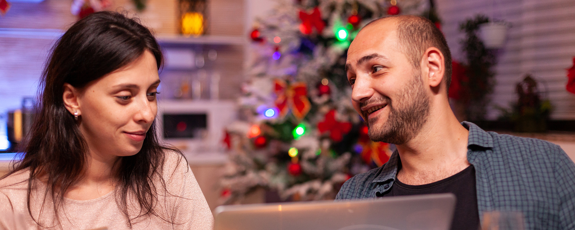 A man and woman sitting at a kitchen table talking about something while looking at their laptop. There is a Christmas tree up in the background.
