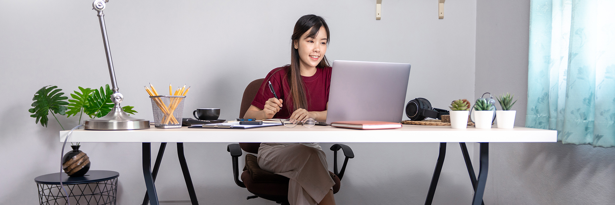 Young woman sitting at her home office desk smiling holding a pen looking at her laptop | How to stay focused WFH